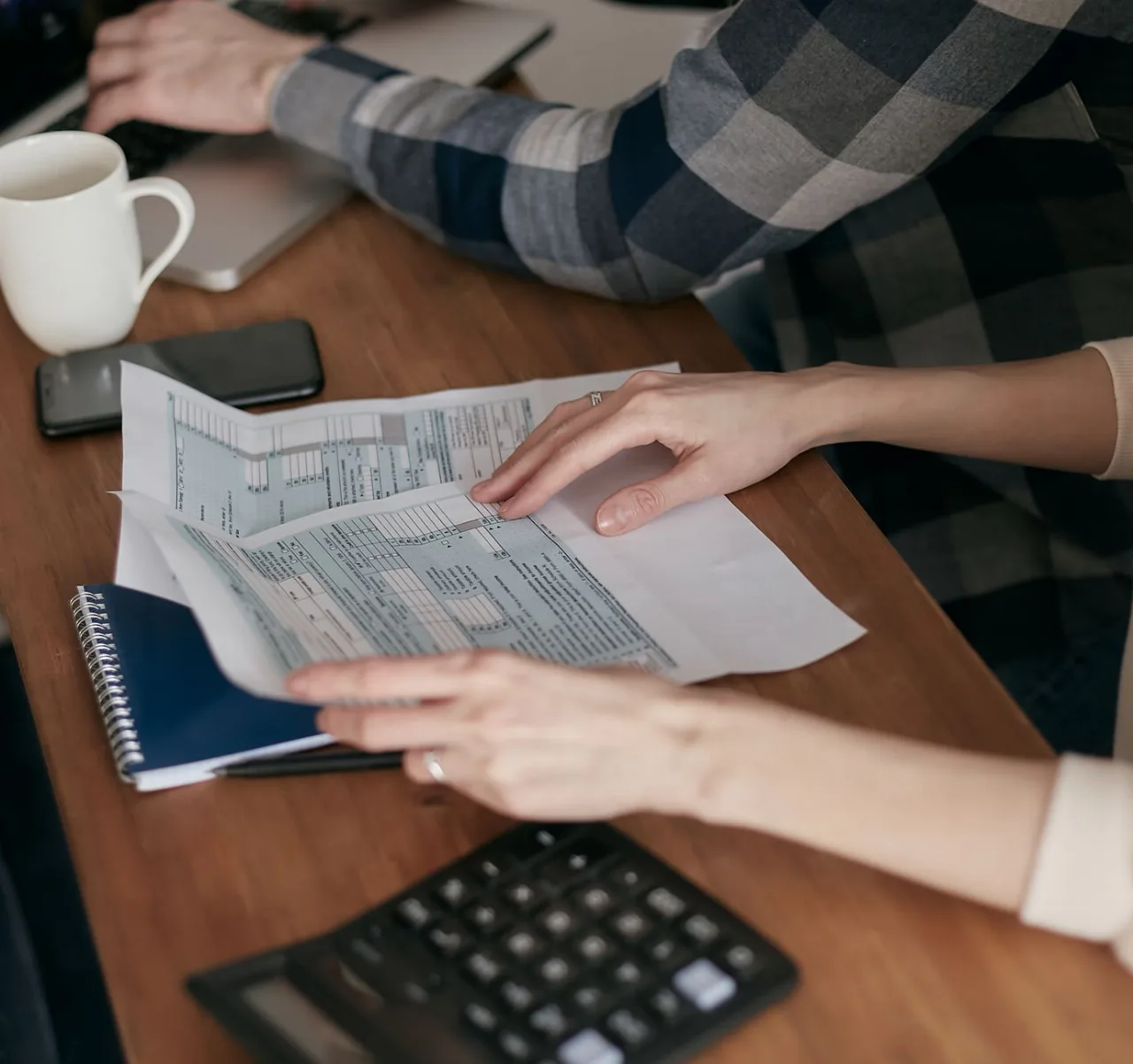 Mãos organizando formulários fiscais sobre uma mesa, ao lado de um caderno e uma xícara de café