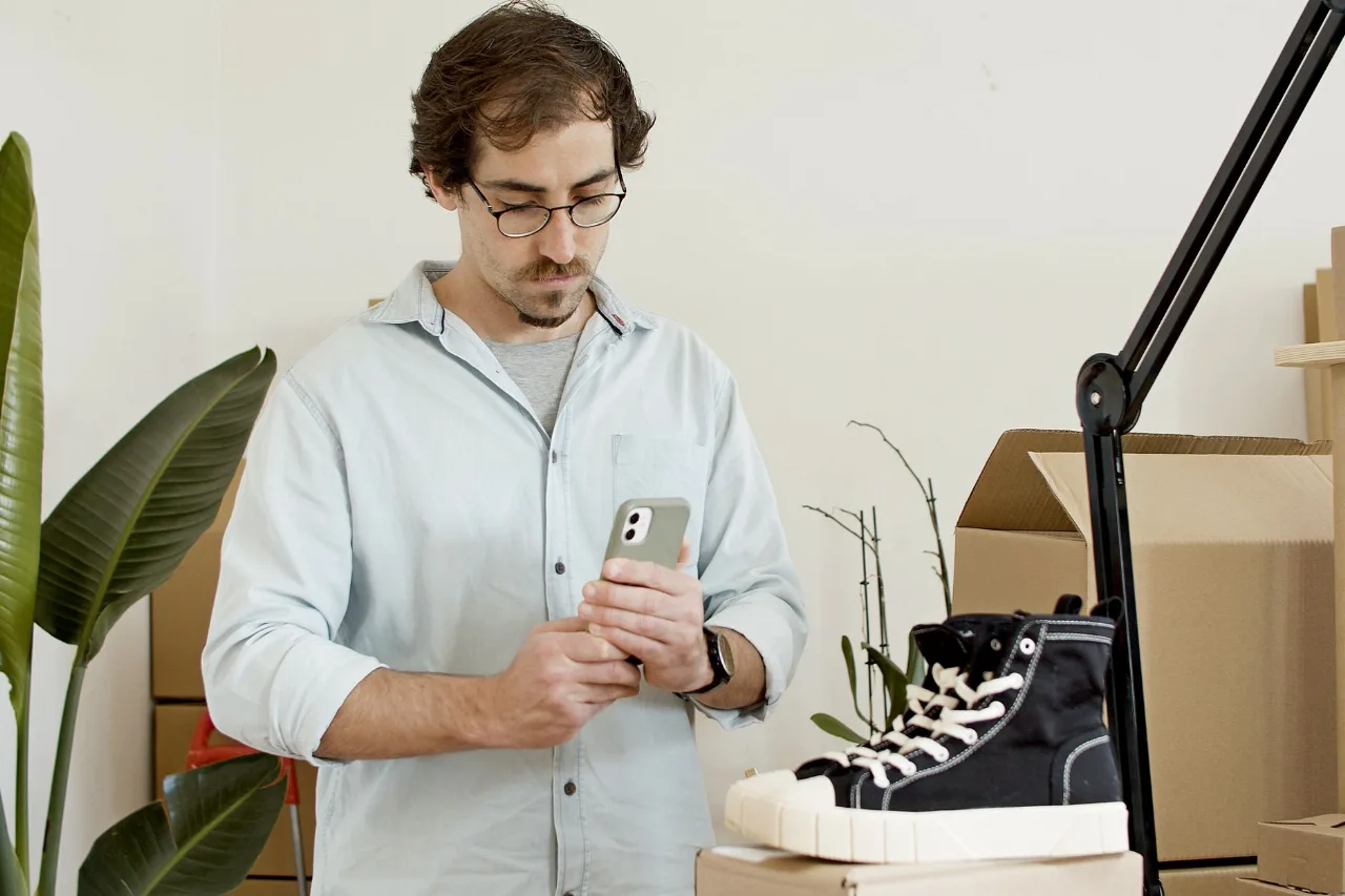 Homem fotografando um tênis com o celular, cercado de caixas de papelão, para anunciar a venda pela internet