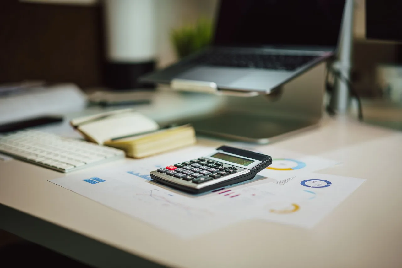 Mesa de trabalho com calculadora, relatórios com gráficos e um notebook ao fundo